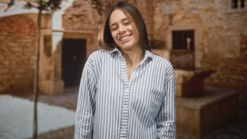 Woman smiling with hands in pockets wearing a striped button shirt, head tilted and eyes soft, standing by a stone fountain beside an old masonry wall in a sunlit building; contentment.