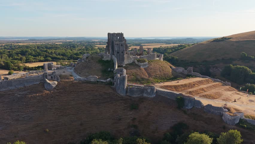 Aerial drone footage of the iconic Corfe Castle in Dorset, England, showcasing the medieval castle ruins, historic village, and rolling Purbeck countryside on a hot summer morning
