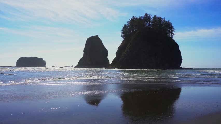 Pan left toward tree topped sea stacks and surf at La Push Second Beach on the Olympic Coast, Washington, with reflections on wet sand. 4K UHD video.