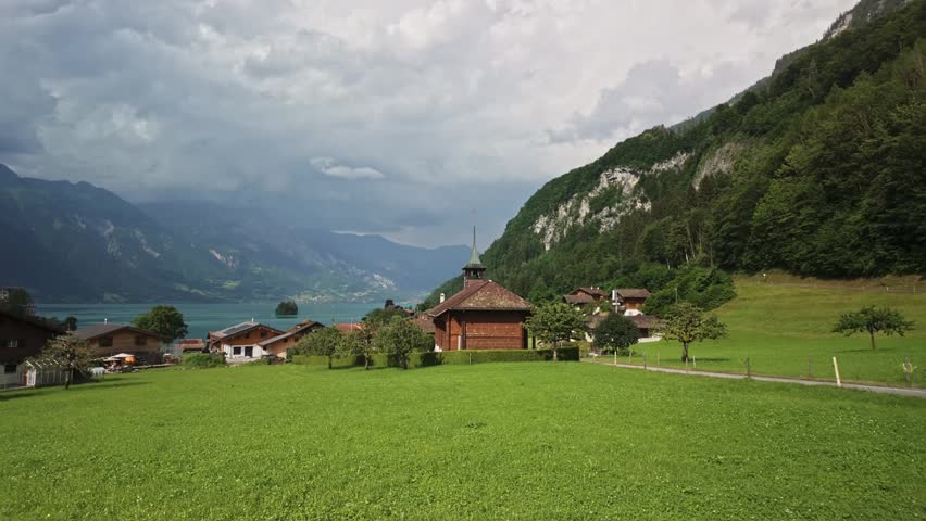 Pan around Iseltwald church amid green alpine meadows above Lake Brienz, with Swiss chalets, forested cliffs, and cloudy Bernese Oberland mountains, Switzerland. 4K UHD video.