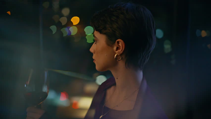 Pensive lady holding red wine glass near window, looking out at blurred city lights night. Close up portrait elegant woman relaxing alone evening, cozy wine bar mood, calm reflection lifestyle.