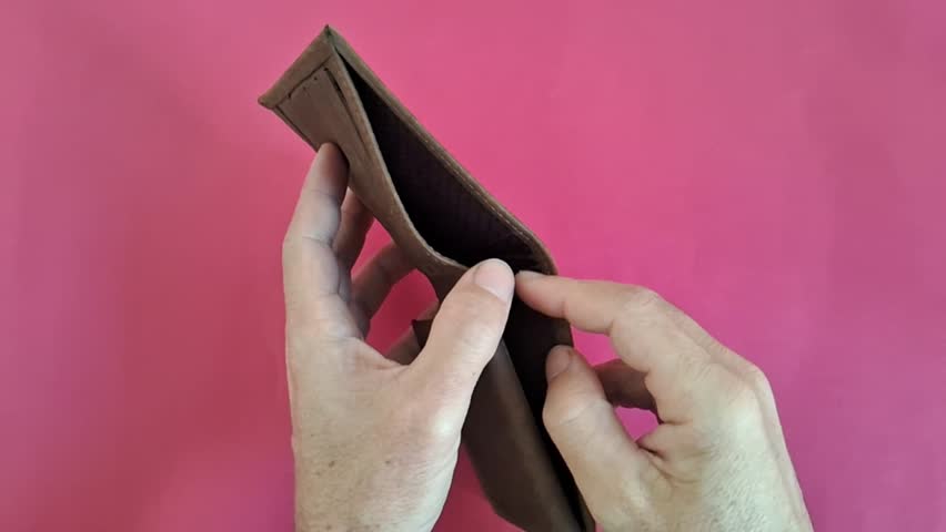 Human hands open an empty brown leather wallet against a pink background illustrating financial hardship and lack of monetary savings.