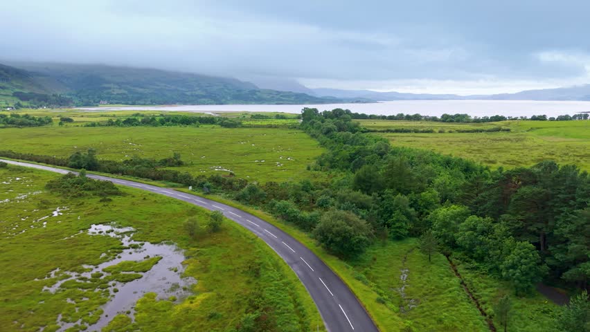 TORRIDON, WESTER ROSS, SCOTLAND - JULY 10, 2026: Aerial tracking view follows a rural road through wet green croftland beside Loch Torridon, with wooded field boundaries and mist-covered hills beyond.