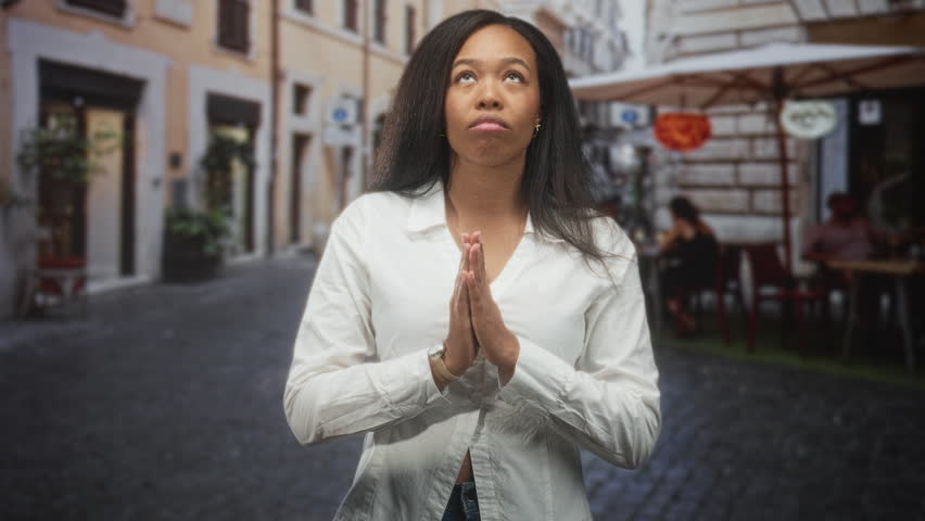 Woman with hands pressed in prayer looking upward on a cobblestone street outside a cafe, wearing a white shirt and jeans; hope prayer.