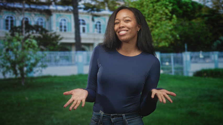 Woman presenting open palms with a slight smirk in front of building on a grassy lawn, wearing navy long sleeve top and jeans; playful confidence.