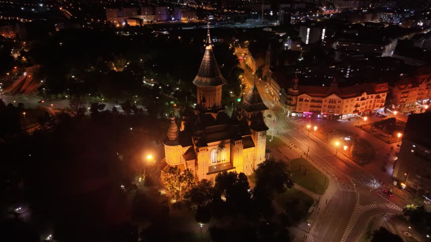 Night aerial view of the illuminated Romanian Orthodox Metropolitan Cathedral in Timișoara, Romania, surrounded by city streets, traffic and urban lights in the historic city centre.