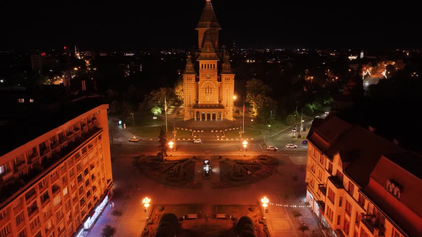 Night aerial view of the illuminated Romanian Orthodox Metropolitan Cathedral in Timișoara, Romania, surrounded by city streets, traffic and urban lights in the historic city centre.