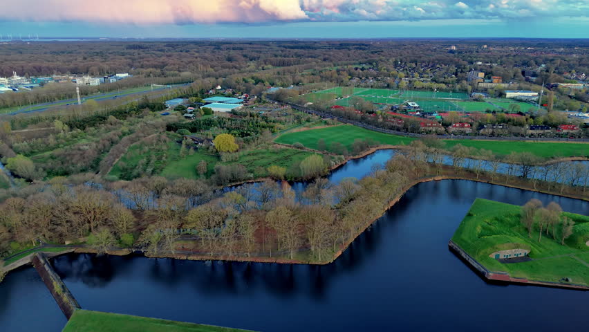 An aerial drone video shot providing a high-angle view over the defensive moat system and green earthen ramparts of the historic star-shaped fortress city of Naarden in North Holland, Netherlands. The video frame highlights the intricate geometric shape of the outer bastions, water canals, and tree-lined defensive walls surrounding the town. In the background, green sports fields, residential areas, and dense forest areas extend toward the horizon under a dramatic twilight sky. Soft sunset light casts delicate pastel pink, orange, and blue tones across the thick clouds and quiet water reflections below. This high-definition video clip showcases historic Dutch fortifications, star-fort architecture, aquatic canal systems, and peaceful countryside surroundings.