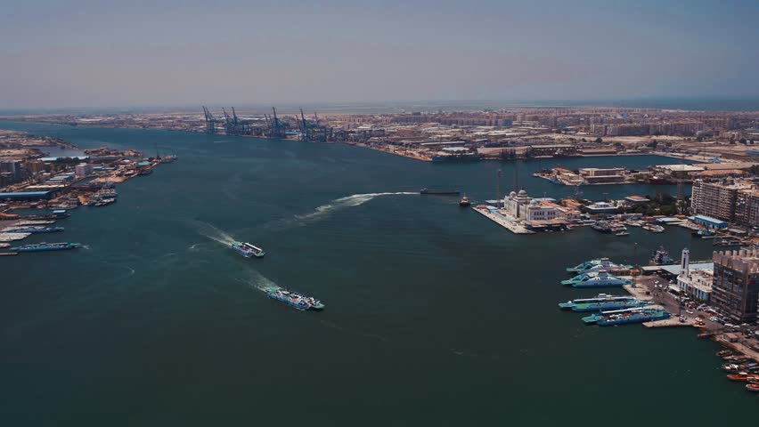 Wide aerial drone view of the Suez Canal in Port Said, Egypt, featuring boats navigating the canal, the Suez Canal Authority waterfront, Port Said harbor, and the surrounding urban skyline at the Mediterranean gateway.