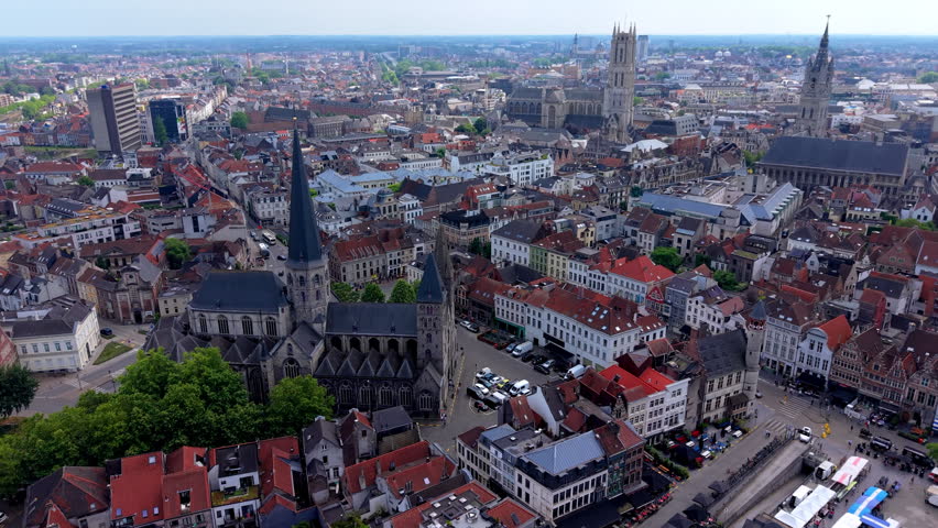 An aerial drone video shot providing a sweeping, high-angle view over the historic heart of Ghent (Gent) in Belgium. The camera movement highlights key landmark architecture, starting with the prominent Saint Nicholas
