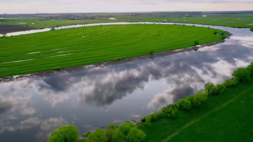 Stunning aerial drone shot of a wide horseshoe-shaped river meander reflecting clouds on its surface, surrounded by vibrant green agricultural fields and country landscape.