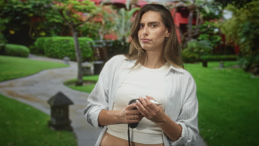 Young hispanic woman holding microphone near mouth, gesturing with open hand on a building courtyard path while singing; joy confidence.