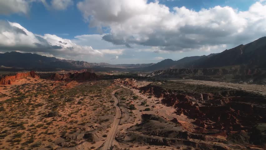 Aerial view of Valles Calchaquies showing the rugged desert landscape with red rock formations, a winding dirt road, and distant mountains in San Carlos, Salta, Argentina.