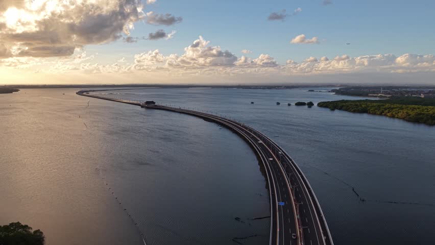 Aerial view of the Mandara Toll Road stretching across the water at sunset with golden light reflecting on the surface and mangrove forests in Kabupaten Badung, Bali, Indonesia.