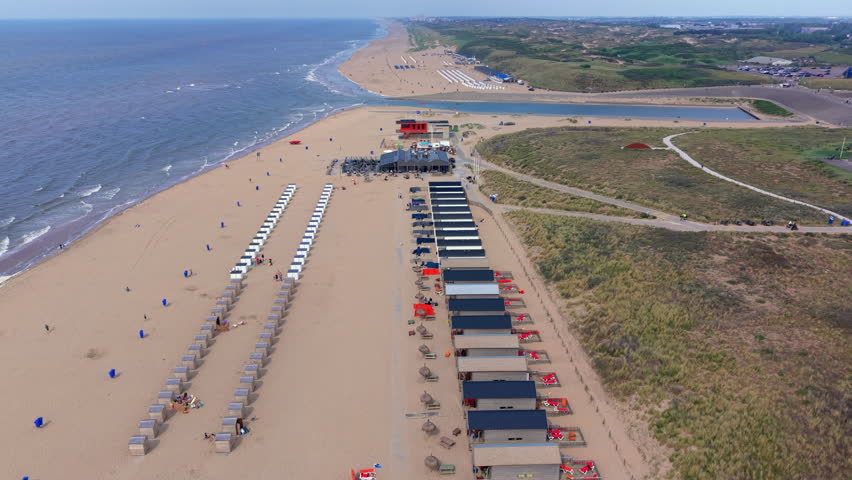 An aerial drone video shot providing a high-angle panoramic view looking down the sandy coastline and coastal dunes of Katwijk aan Zee in the Netherlands. The video frame features a long row of modern beach houses and wooden beach cabins alongside neatly lined-up chairs on the golden sand. In the midground, a seaside beach pavilion sits near a red station building where the Oude Rijn river mouth meets the North Sea behind the dunes. To the right, paved bicycle paths wind through grassy sand dunes, while the blue waters of the sea stretch toward the horizon under a clear summer sky. This high-definition video footage highlights Dutch coastal landscape, seaside tourism, beach resort infrastructure, and peaceful summer vacation scenery.