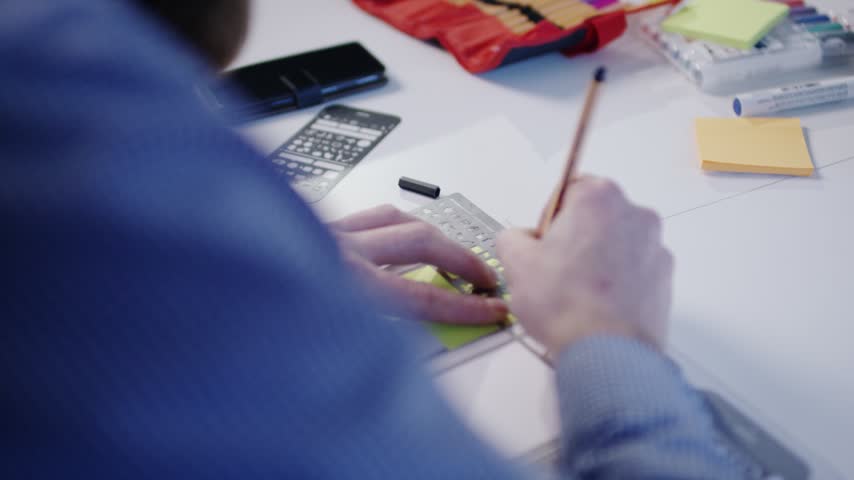 Filmed over the shoulder a person in a blue checkered shirt draws on paper with a wooden pencil and a metal ruler. A case of colored pencils yellow sticky notes a marker and a stencil lie ready on the bright desk. The hand guides the pencil calmly along the ruler in soft daylight. It suits planning and design image films technical explainer videos and education content.