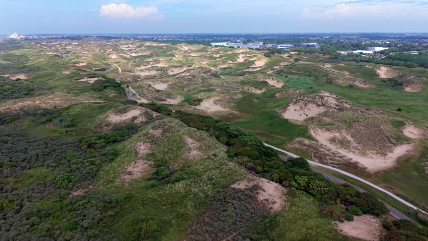An aerial drone video shot providing a high-angle panoramic view over the expansive coastal dune reserve near Katwijk aan Zee in the Netherlands. The video frame features undulating sand dunes covered in marram grass, coastal shrubs, and green vegetation. A paved road and parallel bicycle trail wind gracefully through the dune landscape, leading toward distant commercial and residential buildings under a bright sky with light fluffy clouds. This high-definition video footage highlights Dutch coastal nature, protected environmental reserves, outdoor cycling infrastructure, and scenic European landscapes.