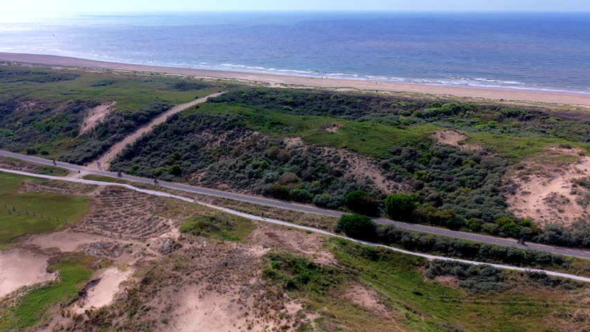 An aerial drone video shot providing a sweeping, high-angle panoramic view over the scenic coastal dune landscape near Katwijk aan Zee in the Netherlands. The video frame showcases a long paved road and an adjacent bicycle path running parallel to the coastline, cutting through dense green coastal vegetation, bushes, and grassy sand dunes. On the upper horizon, a wide sandy beach stretches alongside the sparkling blue waters of the North Sea under clear, bright daylight. Along the path, people can be seen cycling through the protected nature reserve. This high-definition video footage highlights Dutch coastal geography, environmental dune conservation, outdoor cycling routes, and European seaside travel.