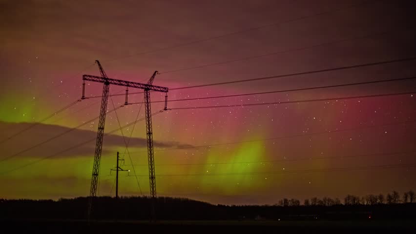 View of vibrant aurora borealis illuminating the night sky behind tall electrical power pylons and transmission lines in the countryside of Latvia.