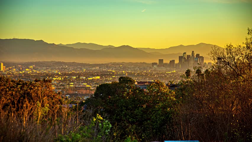 View of the Los Angeles skyline at sunset with distant mountains and foreground trees in Los Angeles, California, United States.