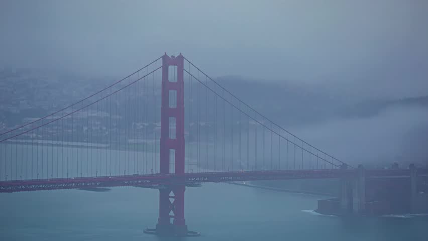View of the iconic Golden Gate Bridge shrouded in thick fog, with the suspension cables and towers visible above the water in San Francisco, California, United States.