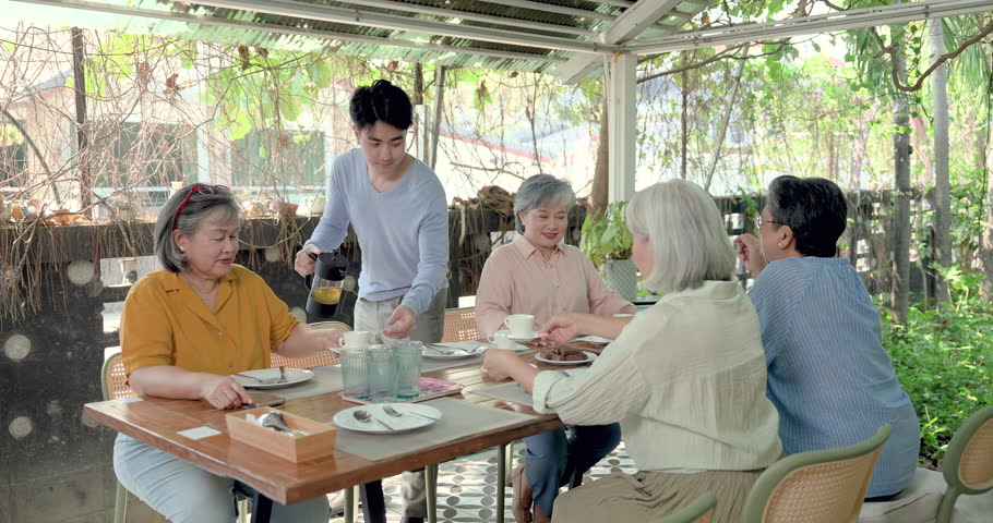 Senior Asian women friend reunion at outdoor restaurant, young man serving coffee at brunch table while retired friends talk together, family gathering, warm hospitality, companionship, relaxed weekend.