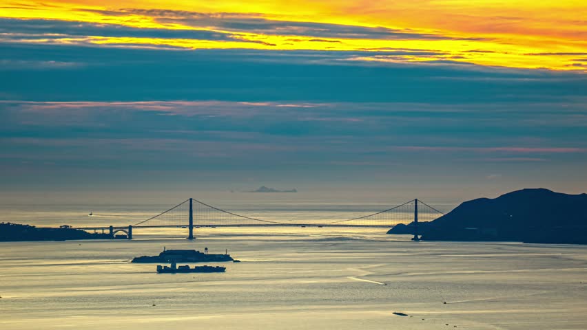 View of the Golden Gate Bridge at sunset with a cargo ship passing near Alcatraz Island under a vibrant sky in San Francisco, California, United States.