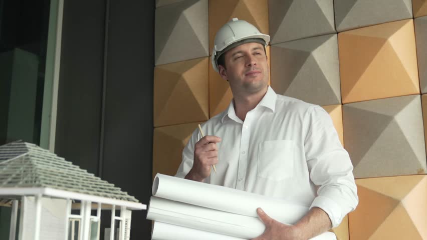 Professional caucasian project manager holding project plan with house model at table. Professional architect engineer wearing safety helmet while standing posing with confident. Manipulator.
