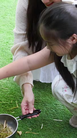 Young girl and woman crouch on grass, feeding a black rabbit from a pan