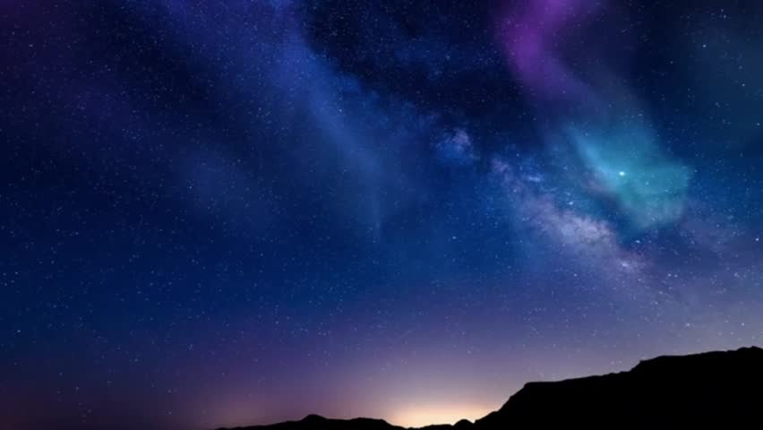 A spectacular night sky view featuring the Milky Way galaxy and shimmering aurora borealis—in shades of purple, light blue, and cyan—glowing above the silhouettes of dark mountains. The sky is filled with millions of brightly shining stars.
