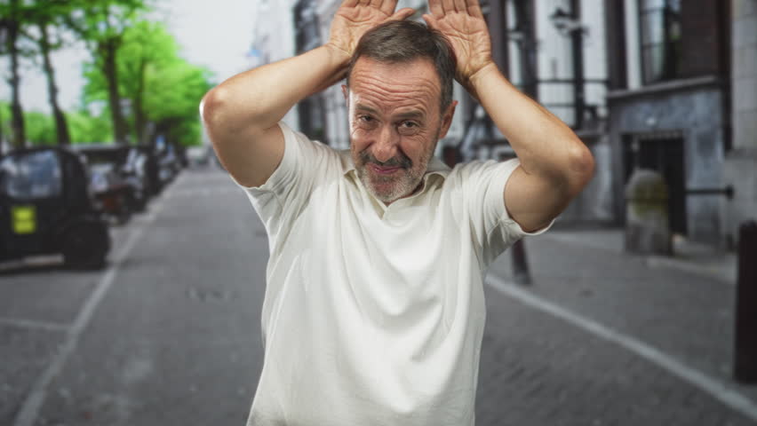 Man with hands on head and open mouth shouting on a cobblestone street in a city, wearing white shirt; playful surprise.