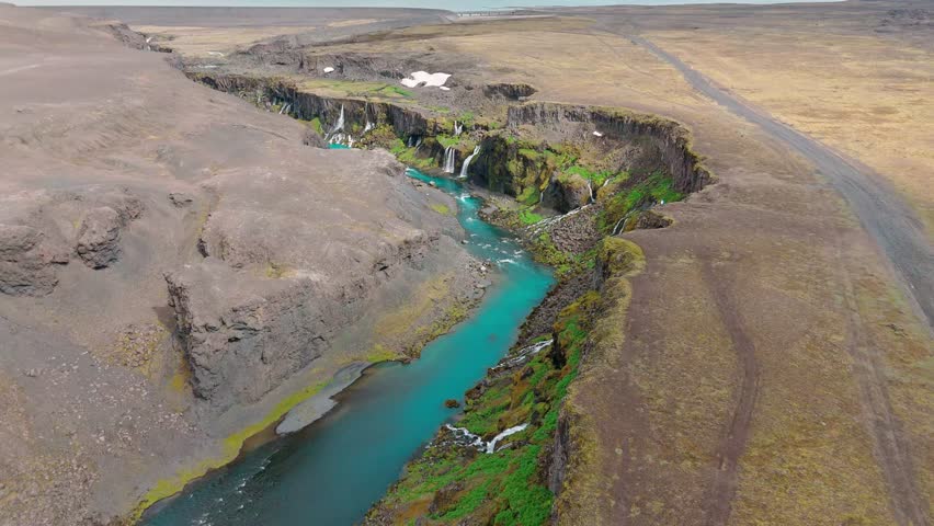 High angle drone shot of the stunning Sigoldugljufur canyon, also known as the Valley of Tears, featuring multiple waterfalls and a vibrant blue river flowing through volcanic terrain.
