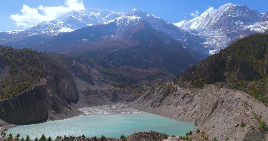 Annapurna Circuit, Manang, Nepal, sideways gliding drone shot over turquoise Gangapurna Lake with snow-capped Himalayan mountain peaks and a glacier in the background on a clear sunny day.