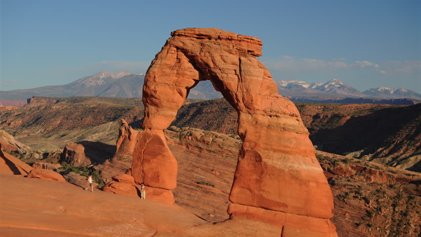 Iconic Delicate Arch stands against blue sky with snow-capped La Sal Mountains in background at Arches National Park Utah. Hikers explore red sandstone formation during golden hour light.