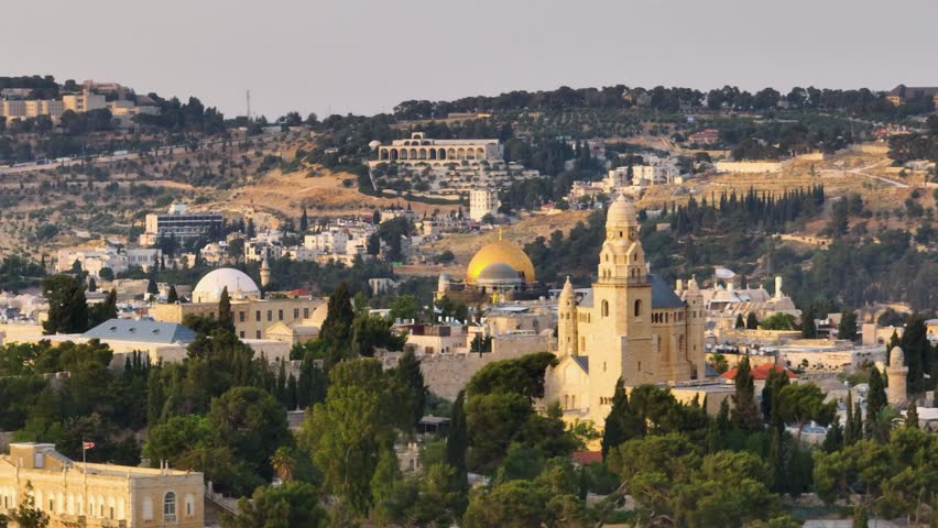 Scenic telephoto shot featuring the iconic Abbey of the Dormition, Dome of the Rock, and Old City skyline bathed in warm evening sunlight.