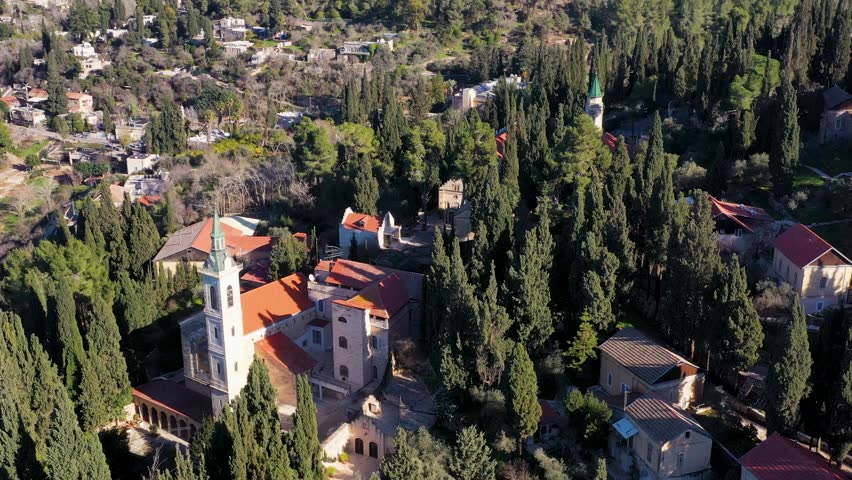 High angle drone shot of historic monastery roofs and church steeple surrounded by lush green cypress forest during bright sunny day in Jerusalem.