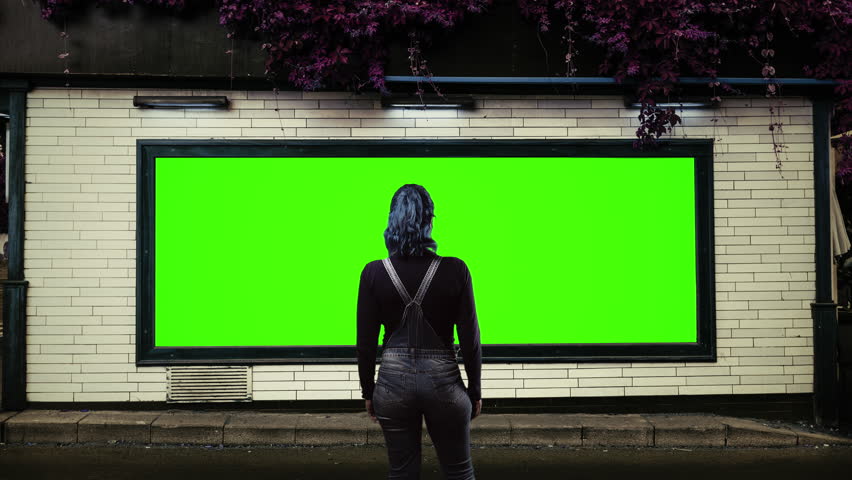 Woman Standing Before Green Screen Billboard in Urban Street, Zoom In. A woman in overalls stands with her back to the camera looking at a large green screen billboard mounted on a tiled urban wall, creating an advertising mockup and creative display opportunity. Zoom In