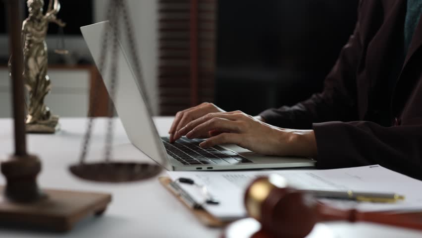 Female lawyer or judge working in a law office, typing on a laptop computer with a gavel, scales of justice, and legal documents on the desk, providing online legal services
