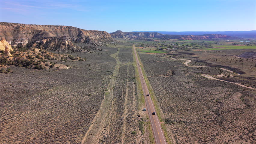 Aerial drone footage captures straight desert highway cutting through arid New Mexico landscape with layered sandstone cliffs, sparse vegetation, and cars traveling under clear blue skies on sunny.