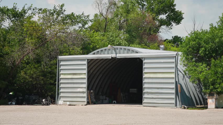 Slow motion shot of a grey steel farm shed with trees swaying in the wind on a rural prairie property during summer