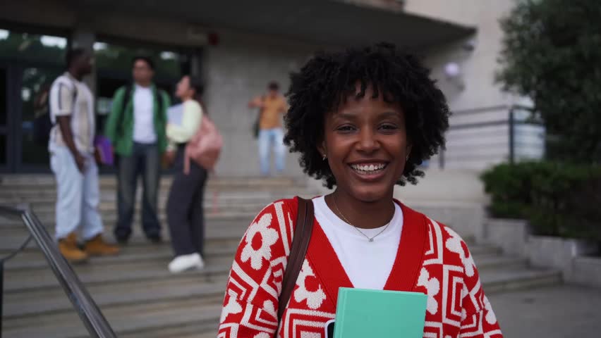 Cheerful young woman with afro hair smiling at camera and laughing in front of a university campus building.