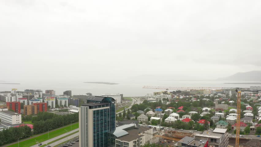 An aerial, high-angle view of Reykjavik, Iceland, on a cloudy day. The cityscape includes modern buildings, a highway, a construction crane, the ocean, and distant mountains.