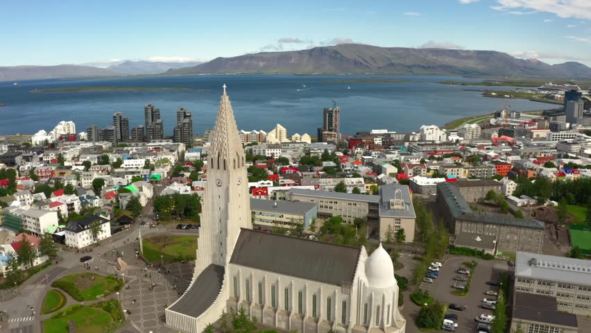 Aerial view flying over Reykjavik, Iceland, featuring the iconic Hallgrímskirkja church, the city center, a bay, and mountains on a sunny day.