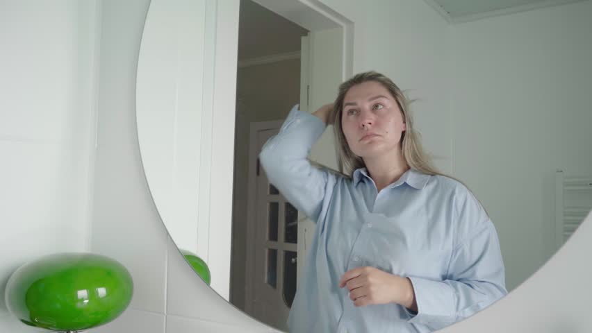 Woman in a light blue button-up smooths her long hair in a round bathroom mirror before an important online job meeting.