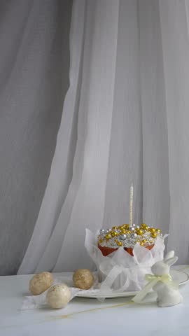 A luxurious Easter cupcake decorated with gold and pearl decorations, a delicate candle, delicate flowers and flowing translucent fabric in a tranquil setting.