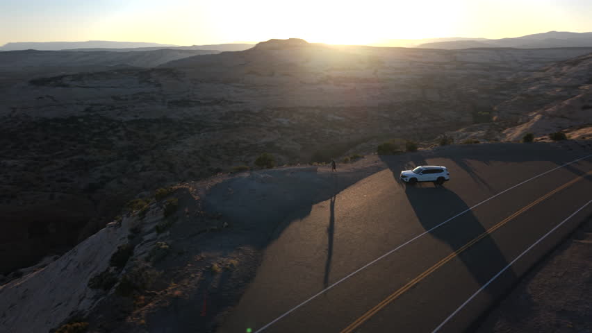 Aerial drone footage captures white SUV parked at scenic overlook along Utah Scenic Byway 12 during golden hour, with dramatic canyon formations, layered rock textures.
