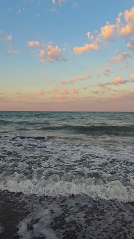 Vertical footage of gentle sea waves crashing on dark sand beach shore during sunset with pink clouds on blue sky background.
