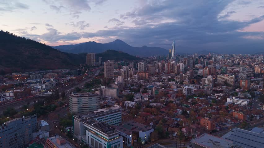 Panning shot over a hillside neighborhood revealing a wide city skyline with distant skyscrapers and mountains at dusk. Santiago, Chile