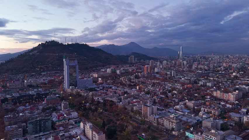 Scenic overview of Cerro San Cristobal hill towering above a dense city neighborhood with distant skyscrapers and mountains at dusk. Santiago, Chile