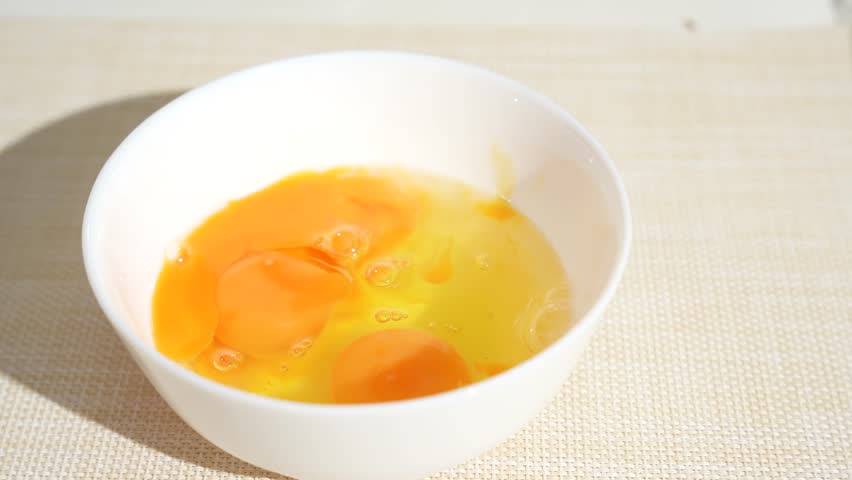 Two raw egg yolks sit in a white bowl as a spoon moves through them. Bright natural light highlights the fresh chicken eggs.